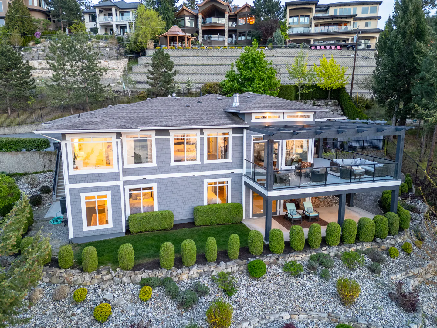 Two-story gray house with a dark gray roof, a patio with furniture, and green bushes in the front yard. Other houses are on a hill in the background.