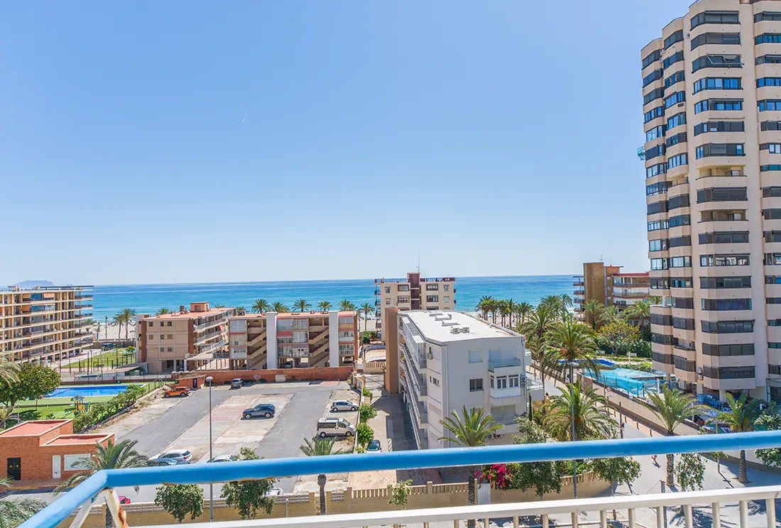 View from a balcony overlooking a coastal city with a beach, blue ocean, palm trees, and tall buildings under a clear blue sky.