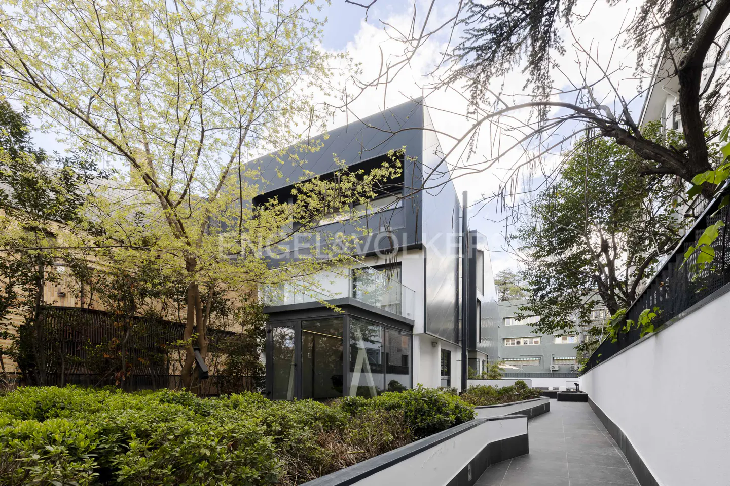 Modern white and black house with a glass-enclosed ground floor, surrounded by lush green trees and a paved walkway.