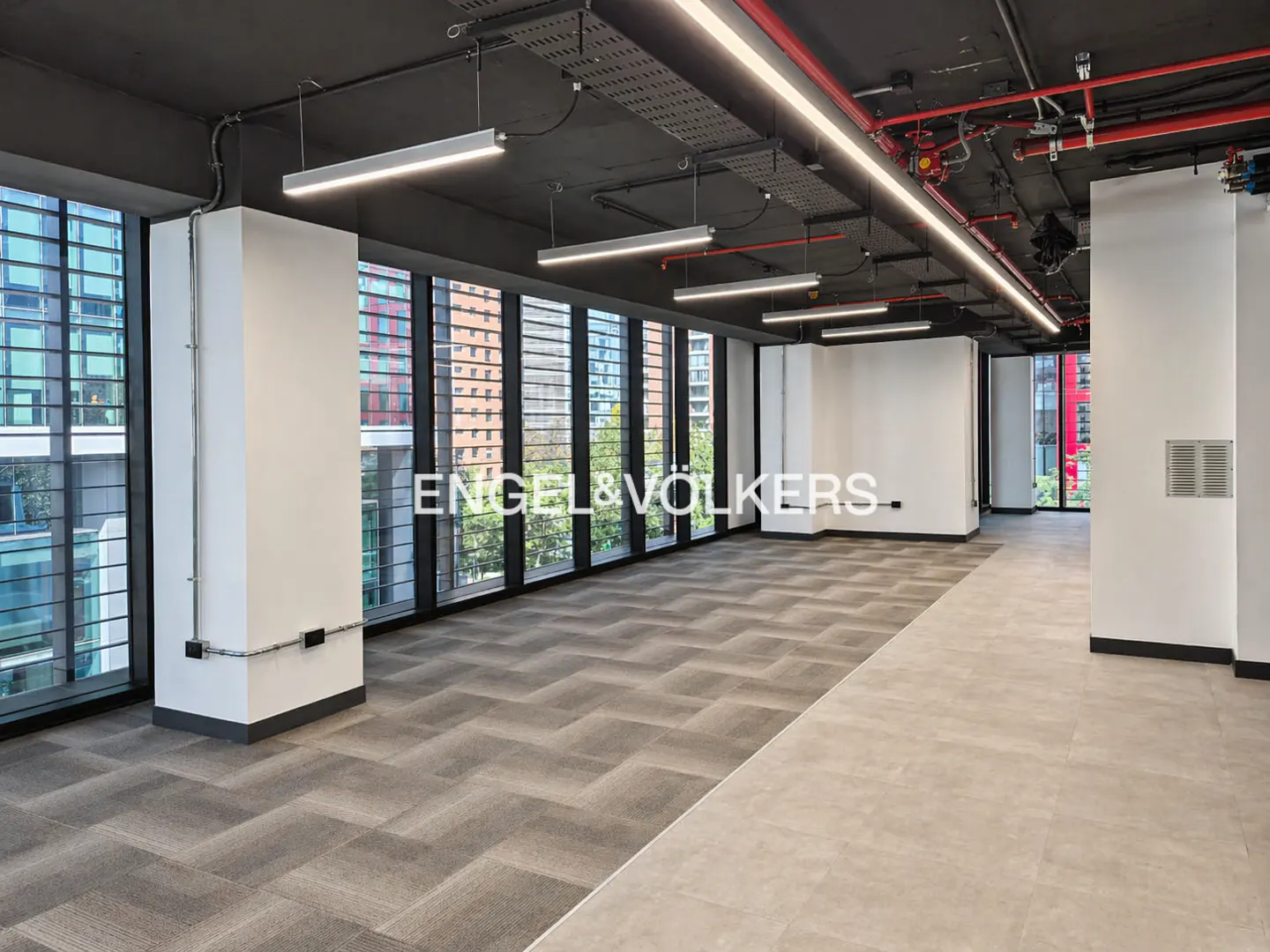 Empty office space with large windows, gray carpet, and white walls. Overhead lighting and exposed ceiling pipes. Engel & Völkers logo visible.