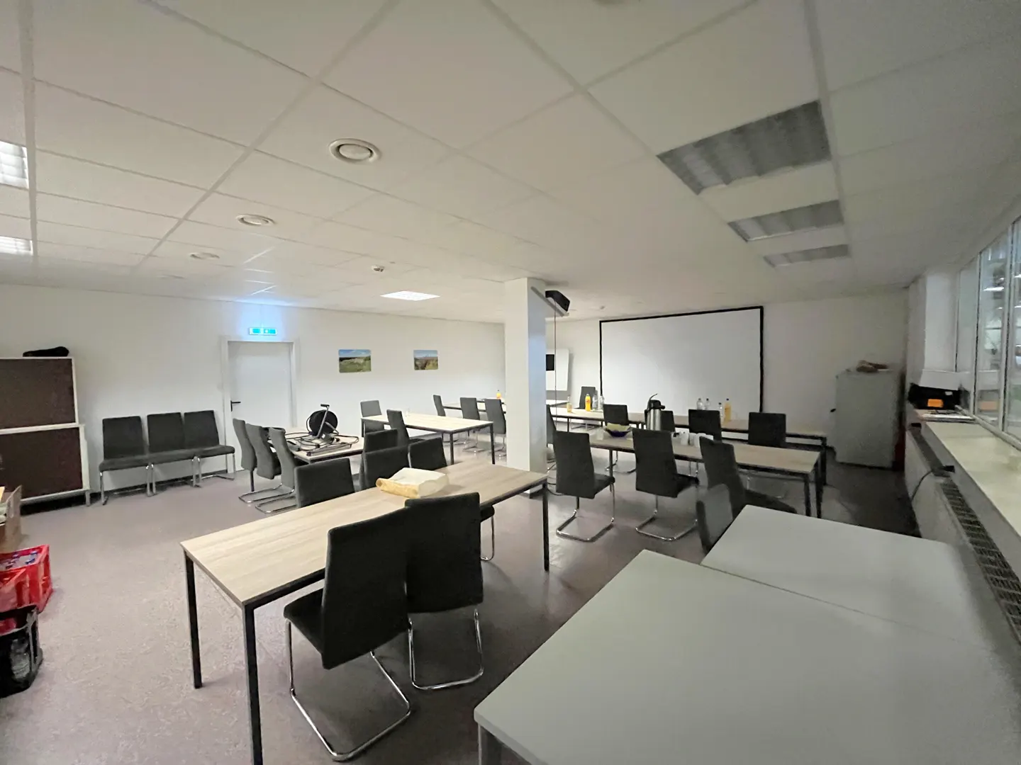A bright, spacious meeting room with rows of light-wood tables, black chairs, a projector screen, and white walls.