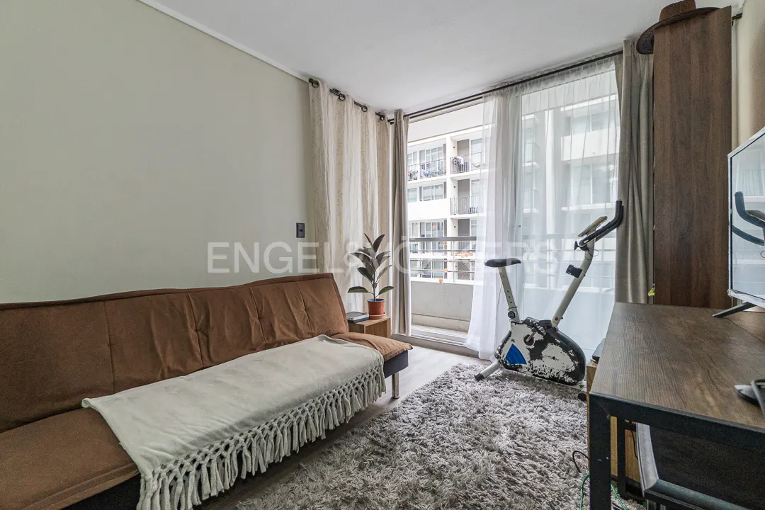A room with a brown sofa, a white fringe throw, a stationary bike, a grey rug, and a balcony view of an apartment building.