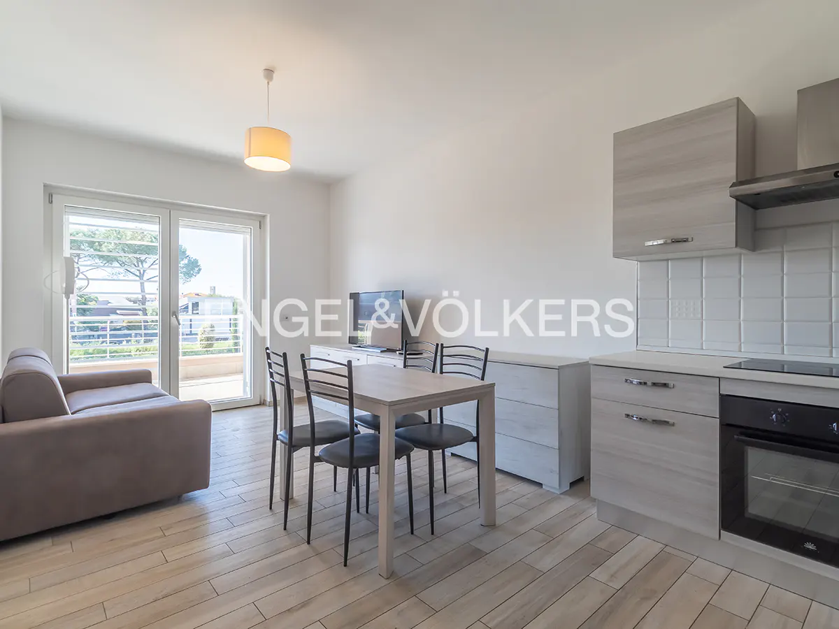 Bright apartment interior with a gray sofa, dining table, and kitchen area. Balcony doors open to a view of trees and buildings.