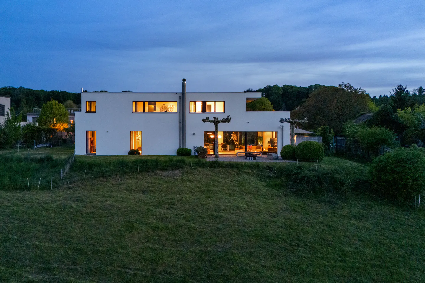 Modern white house on a grassy hill at dusk. Lights are on inside, visible through windows and sliding glass doors.