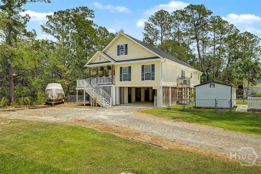 Two-story yellow house on stilts with black shutters, white stairs, and a boat parked in the gravel driveway.