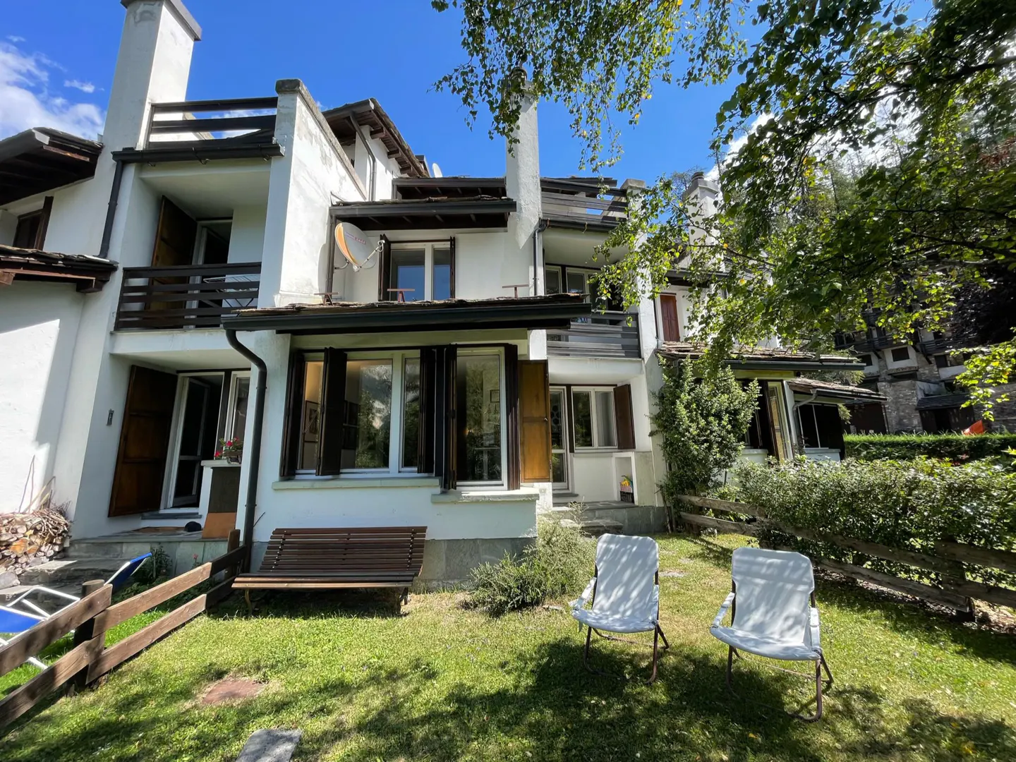 Exterior of a white multi-story house with dark shutters, a bench, and two white chairs on a green lawn.