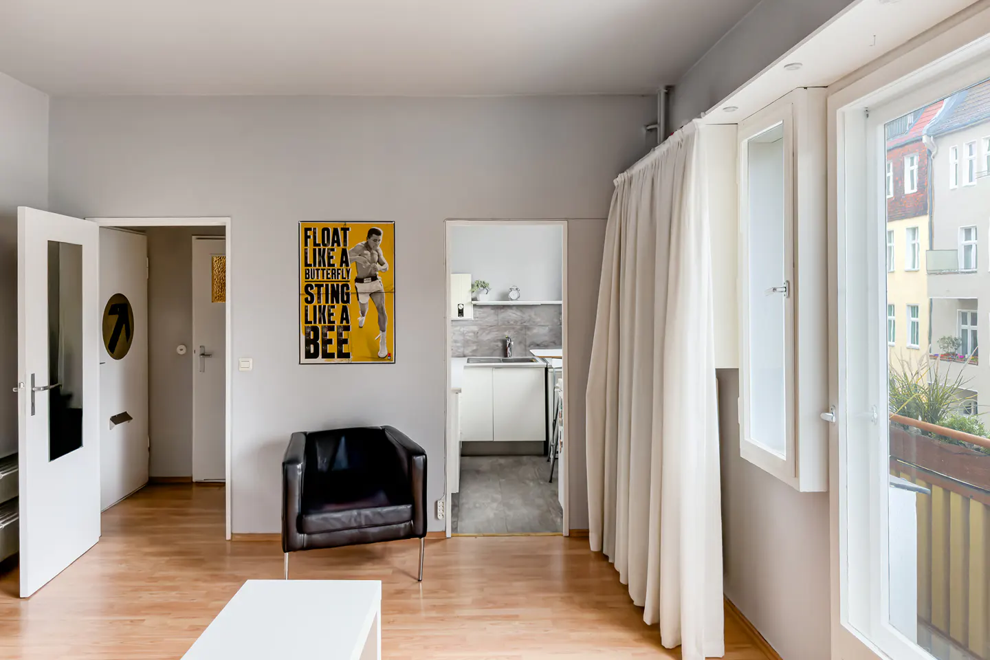 A gray room with wood floors, white trim, and a black leather chair. A Muhammad Ali poster hangs on the wall. A kitchen is visible through a doorway.