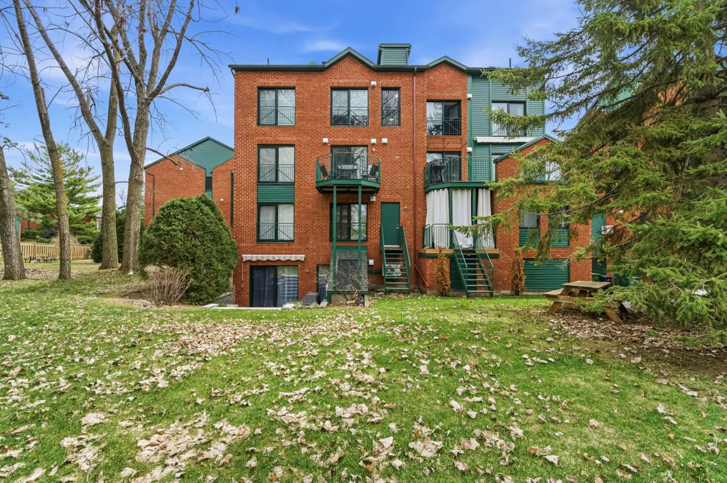 Back view of a red brick apartment building with green trim and balconies, surrounded by a grassy yard with fallen leaves.