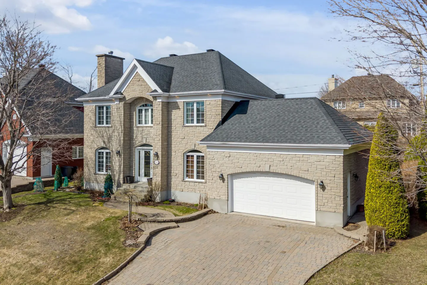 Two-story stone house with a gray roof and a two-car garage, with a brick driveway and a lawn.