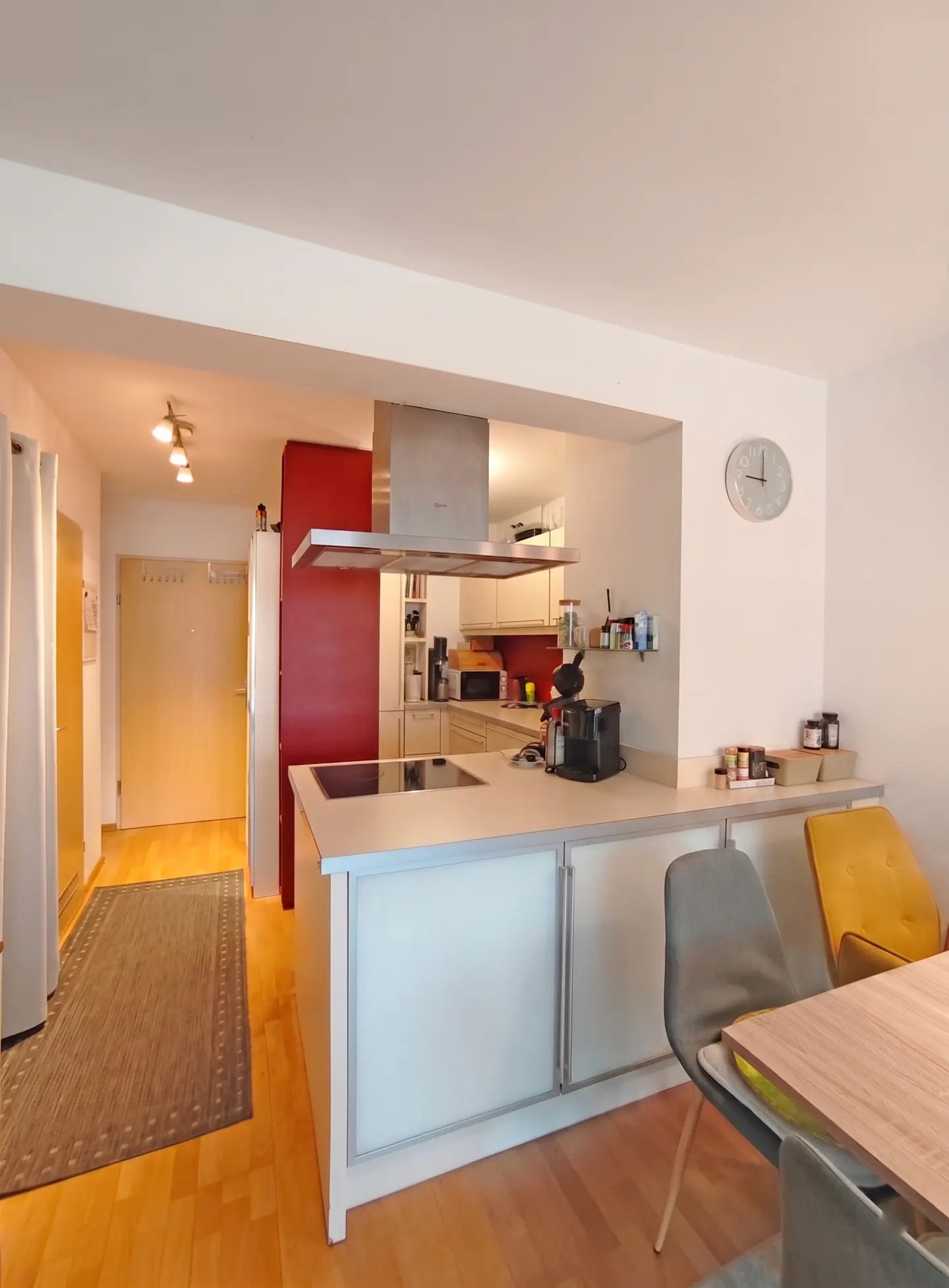 A bright, modern kitchen with a central island, stainless steel hood, and a red accent wall. A dining table with chairs is visible in the foreground.