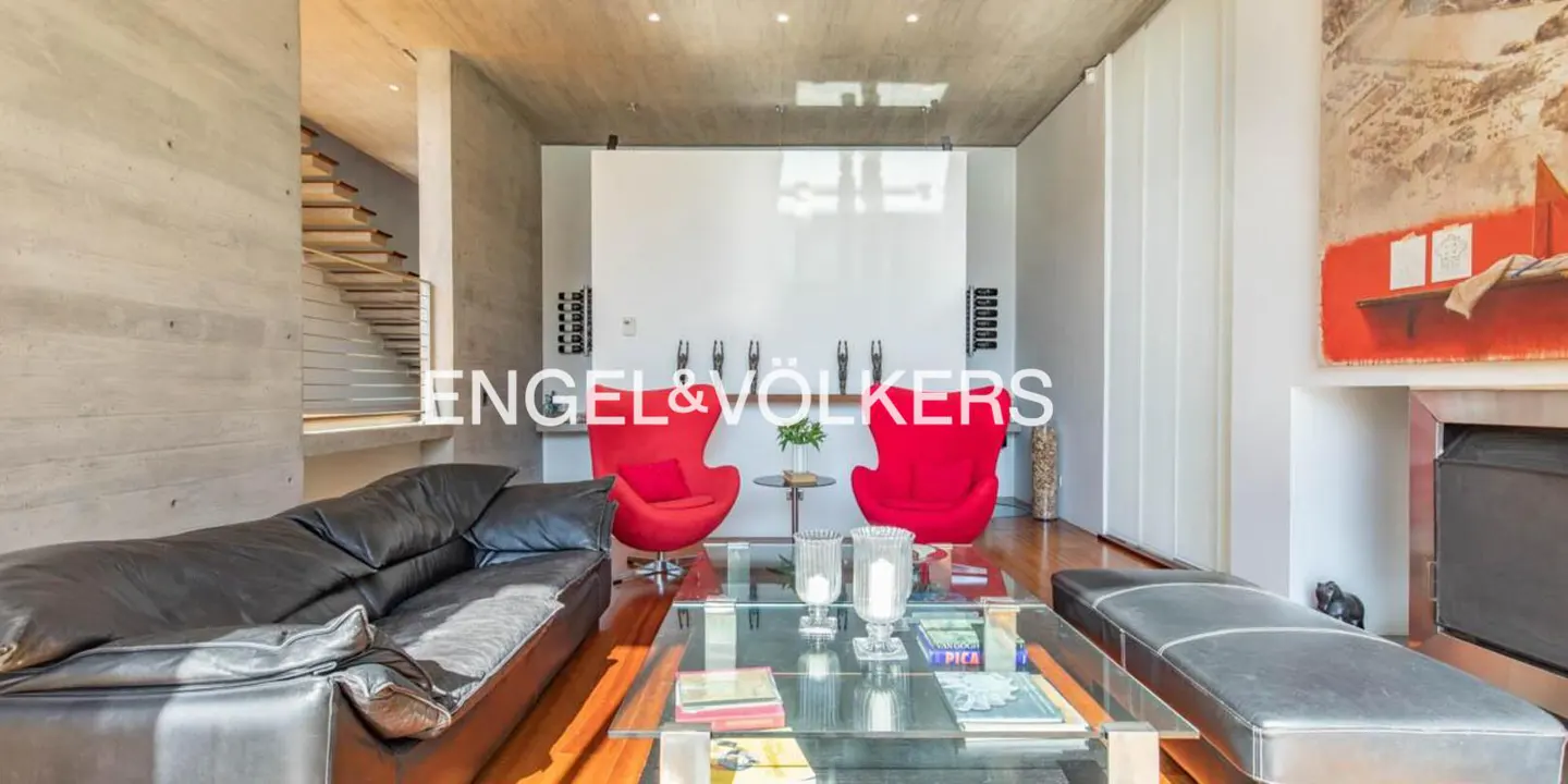 Living room with concrete walls, hardwood floors, black leather sofa, two red chairs, and a glass coffee table. Stairs are visible on the left.