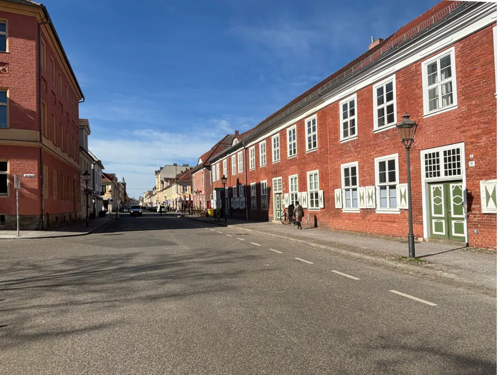 Street view of red brick buildings with white windows and green doors under a blue sky. A car is seen in the distance.