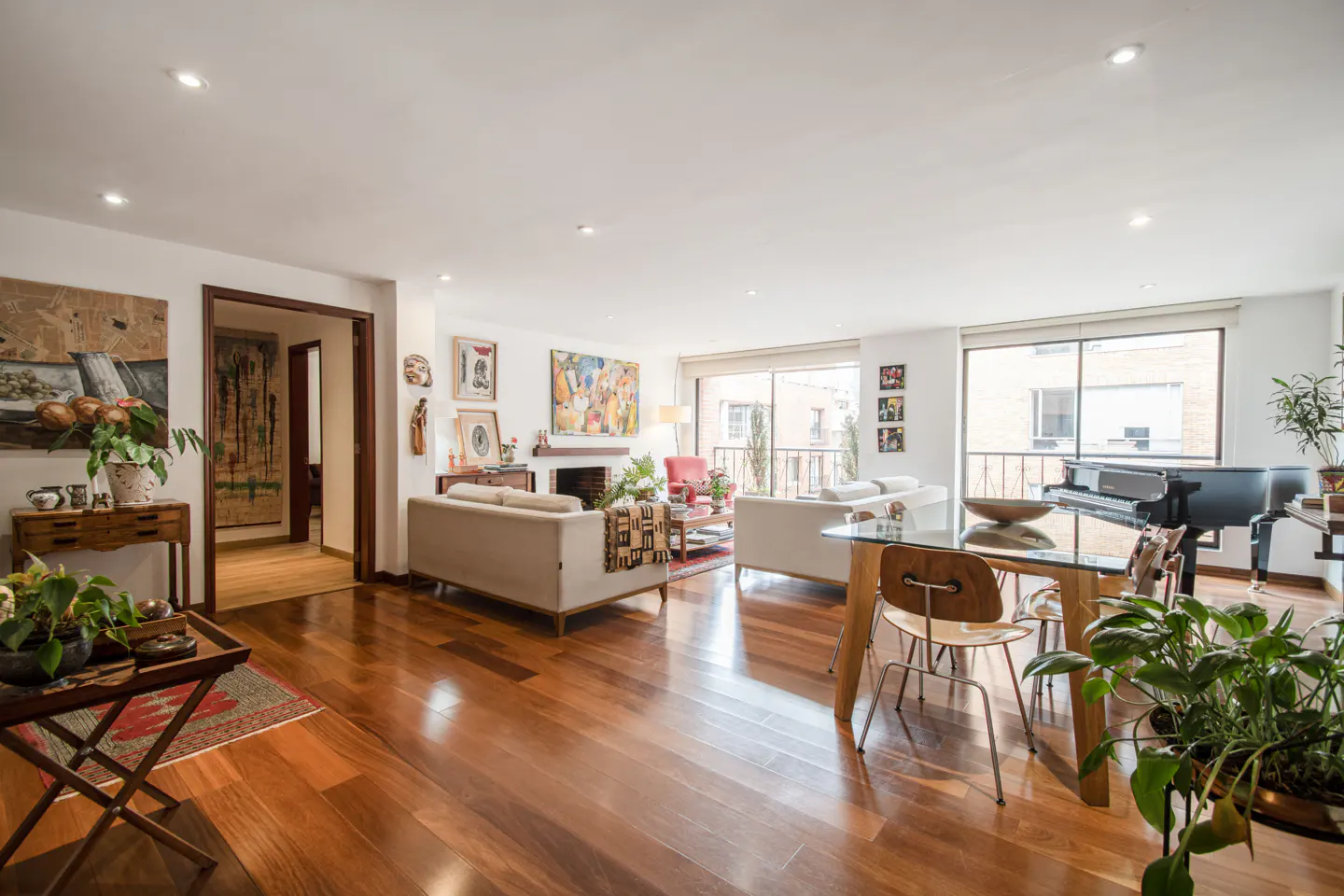 Bright, open living room with hardwood floors, white sofas, a glass dining table, and a black piano by a large window. Art adorns the walls.