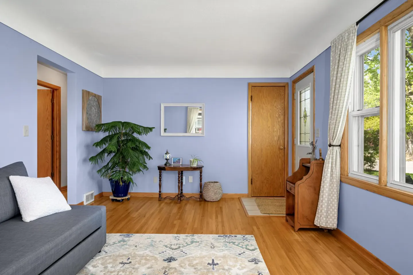 Living room with light blue walls, hardwood floors, and a gray sofa. A large window with curtains lets in natural light. A wooden door and table add warmth.