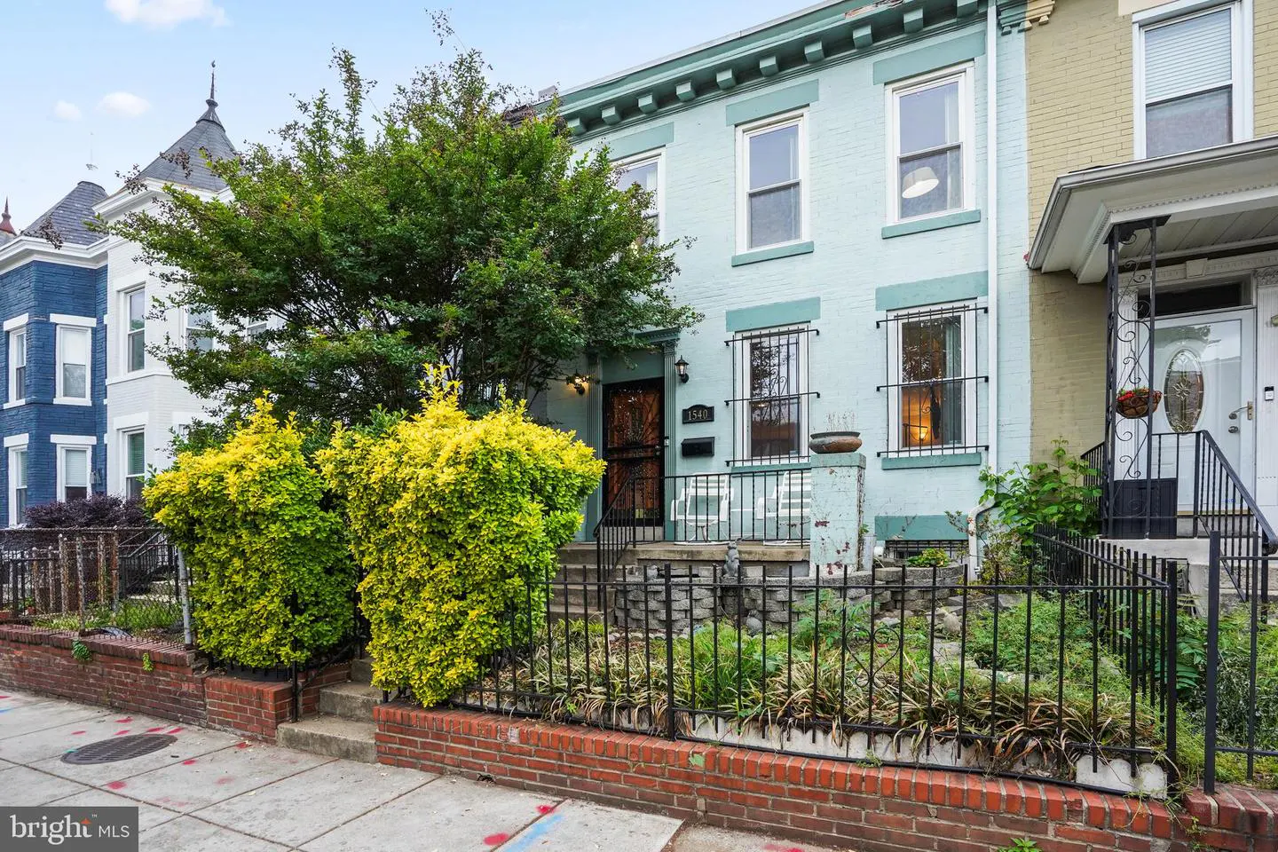 A two-story, light blue row house with a black iron fence and green bushes in front.