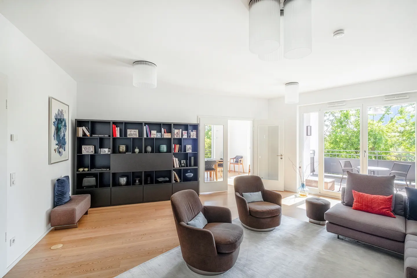 Bright living room with hardwood floors, a large black bookshelf, two brown armchairs, and a sofa with a red pillow.