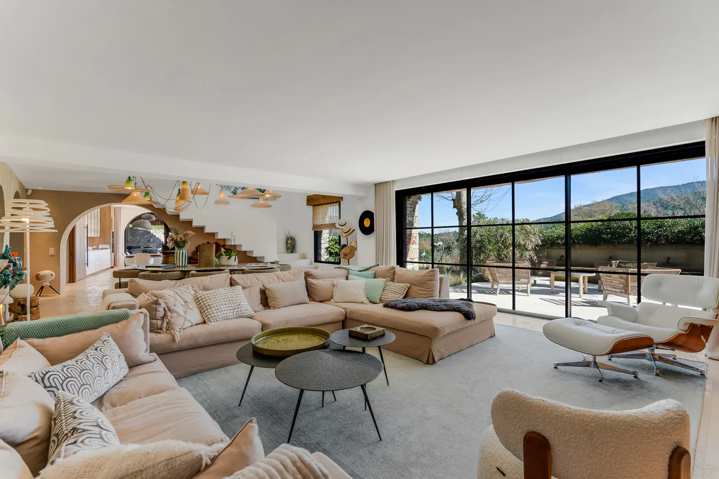 Spacious living room with beige sofas, round tables, and large windows overlooking a patio and greenery.