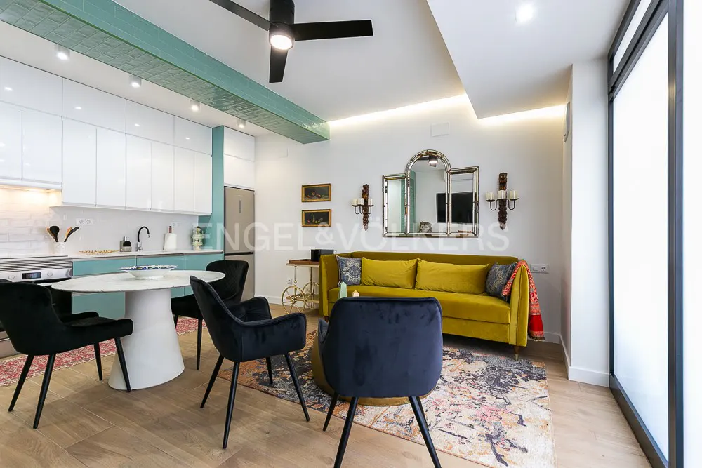 Modern open-plan living area with a white kitchen, teal accents, a mustard sofa, and black dining chairs on a patterned rug.