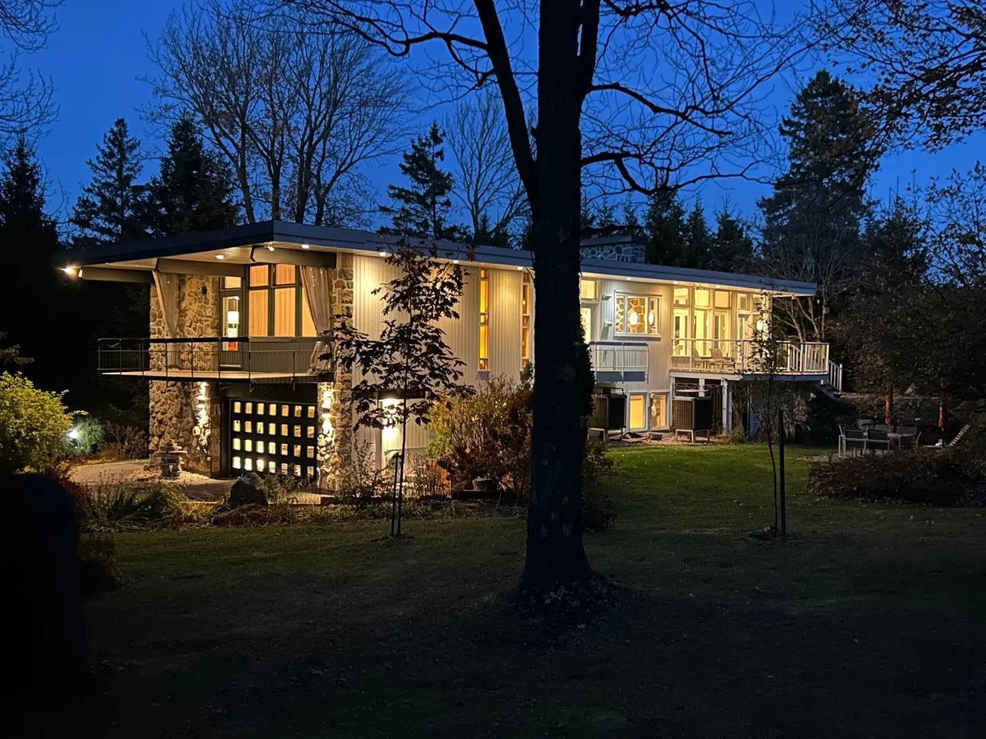 Two-story house with stone and light siding, lit up at dusk. Balconies, garage with square windows, and trees surround the property.