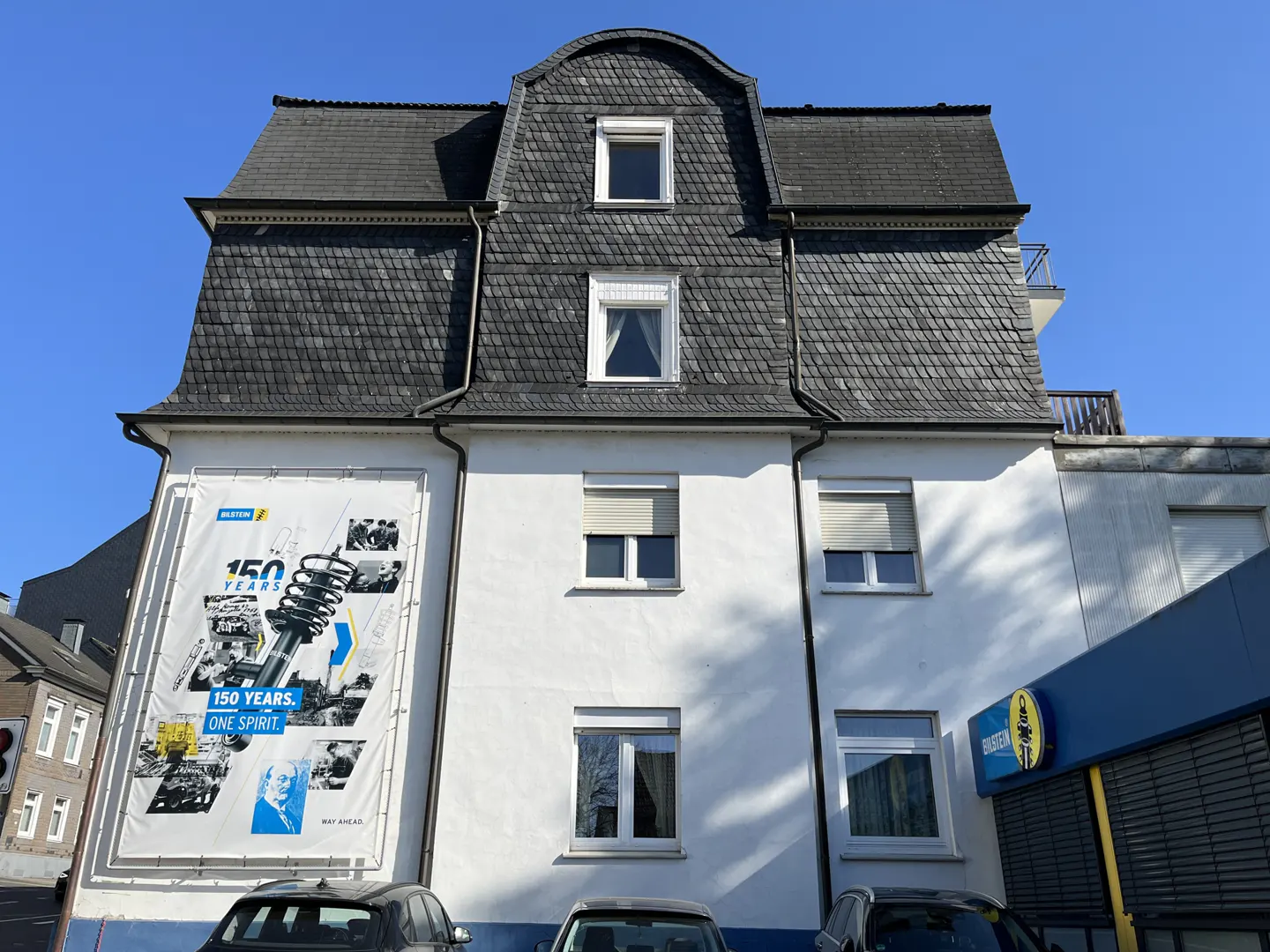 A white building with a dark gray slate roof and several windows. A large Bilstein banner hangs on the left side of the building.