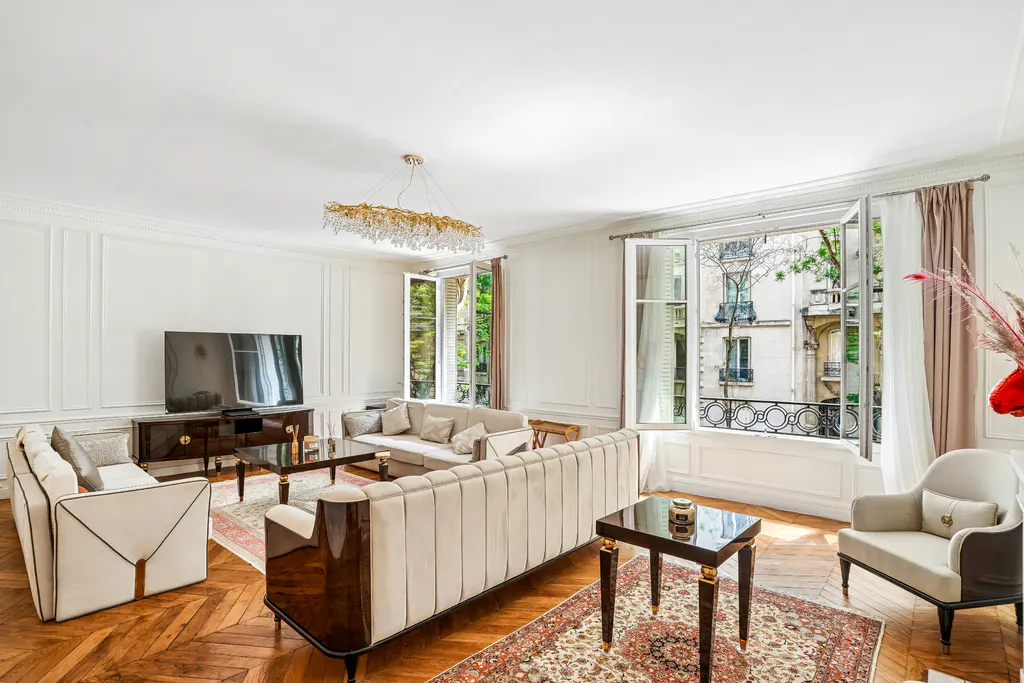 Bright living room with white walls, herringbone wood floors, and two beige sofas. Large windows overlook a Parisian street.