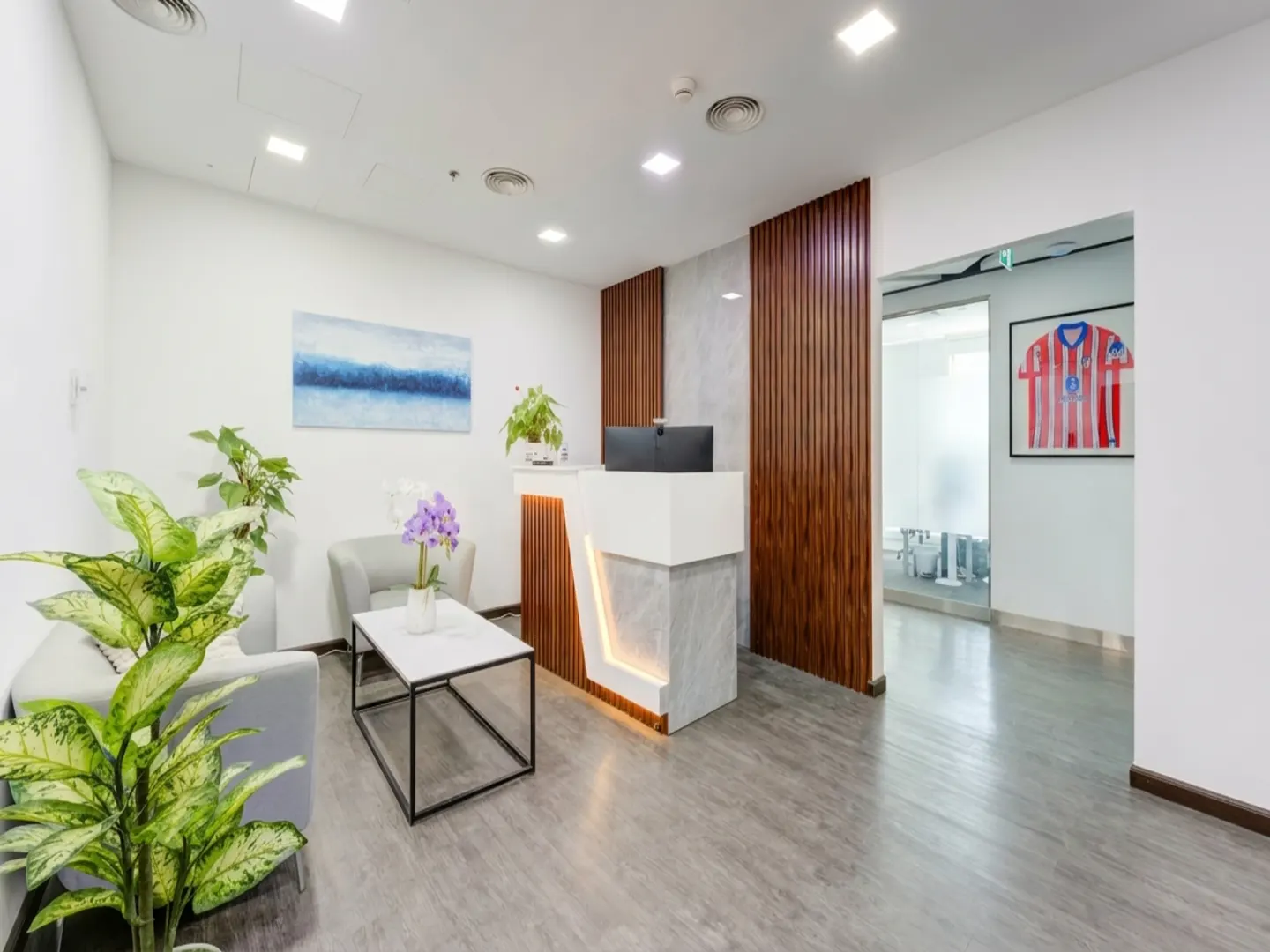 Modern office reception area with white walls, gray floors, a white reception desk, and a seating area with plants.