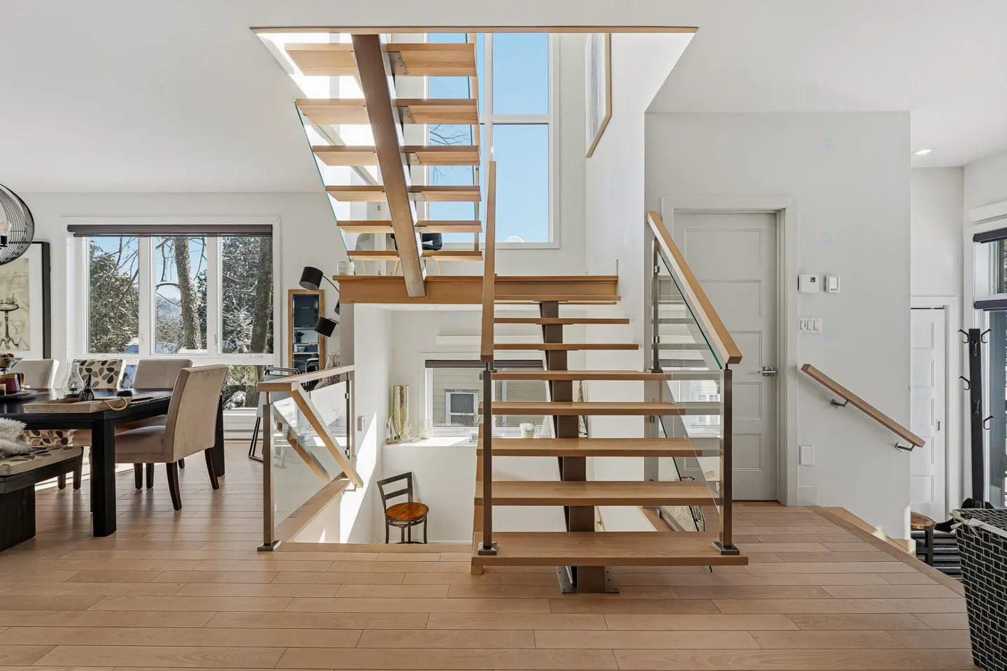 Modern open-concept living area featuring a floating wooden staircase with glass railings and a large window.