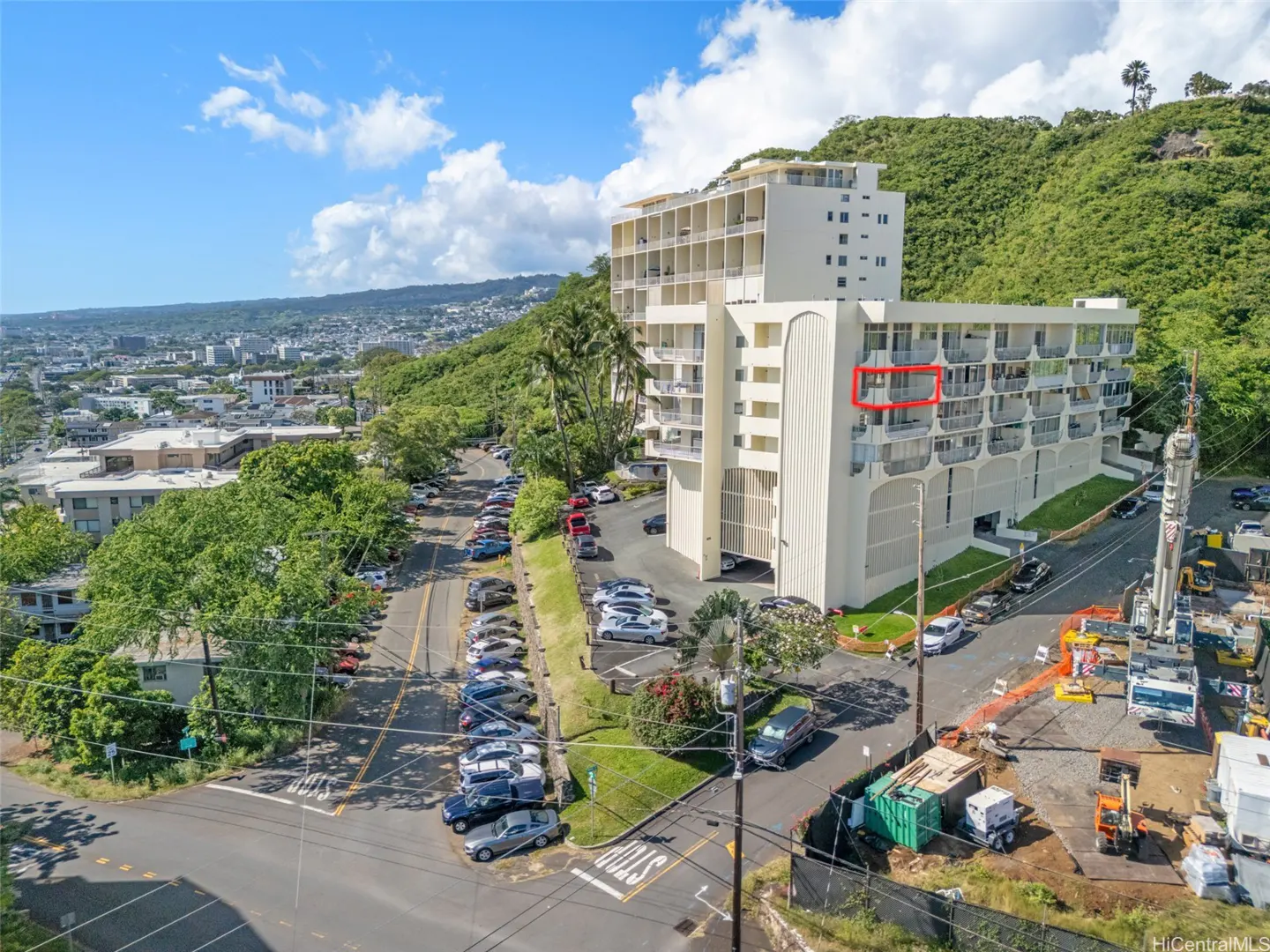 Aerial view of a white condo building with a red box highlighting a unit, surrounded by lush greenery and city views.
