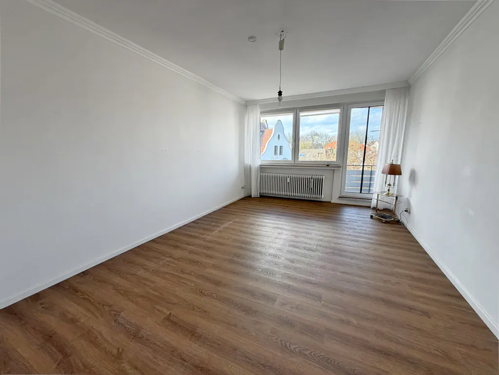 Bright, empty room with wood floors, white walls, and large windows. A radiator sits under the windows, and a lamp is on a table to the right.
