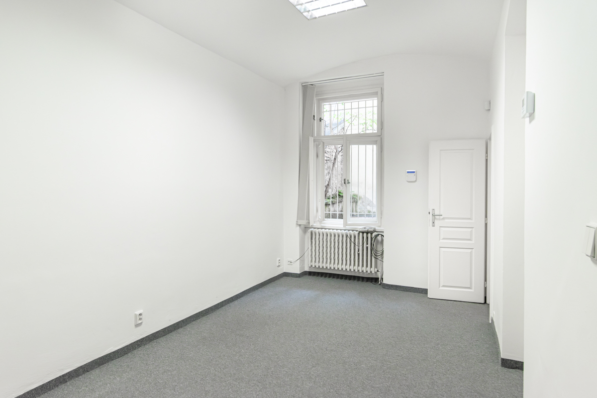 Bright, empty room with white walls, gray carpet, and a window with bars. A white door is on the right.