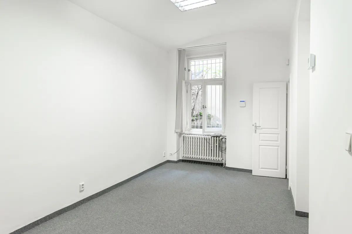 Bright, empty room with white walls, gray carpet, and a window with bars. A white door is on the right.