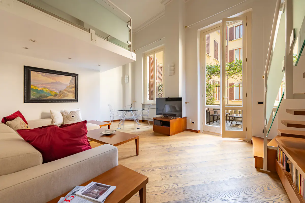 Bright living room with a beige sofa, red pillows, wood floors, and a glass table. French doors open to a courtyard. A painting hangs above the sofa.