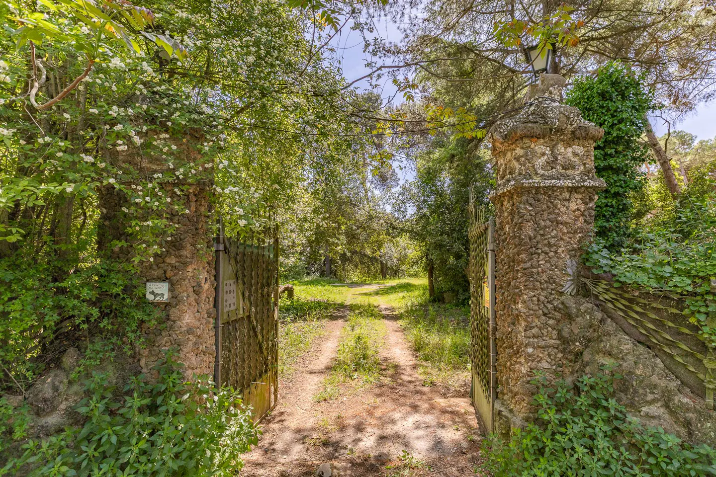 Open wrought iron gate with stone pillars leads to a grassy path lined with trees.