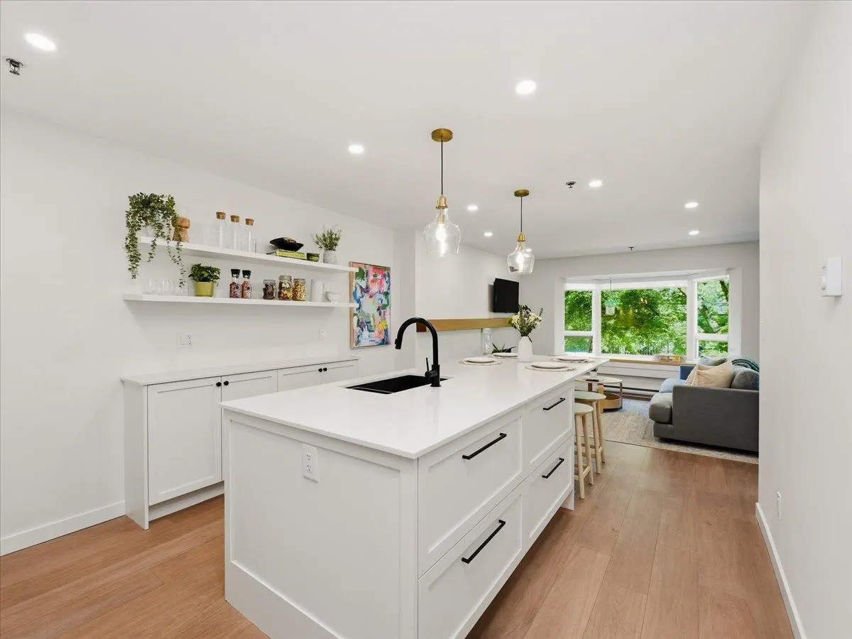 Bright, open-concept kitchen with white cabinets, island, and wood floors. Shelves display plants and jars. Living room visible in the background.