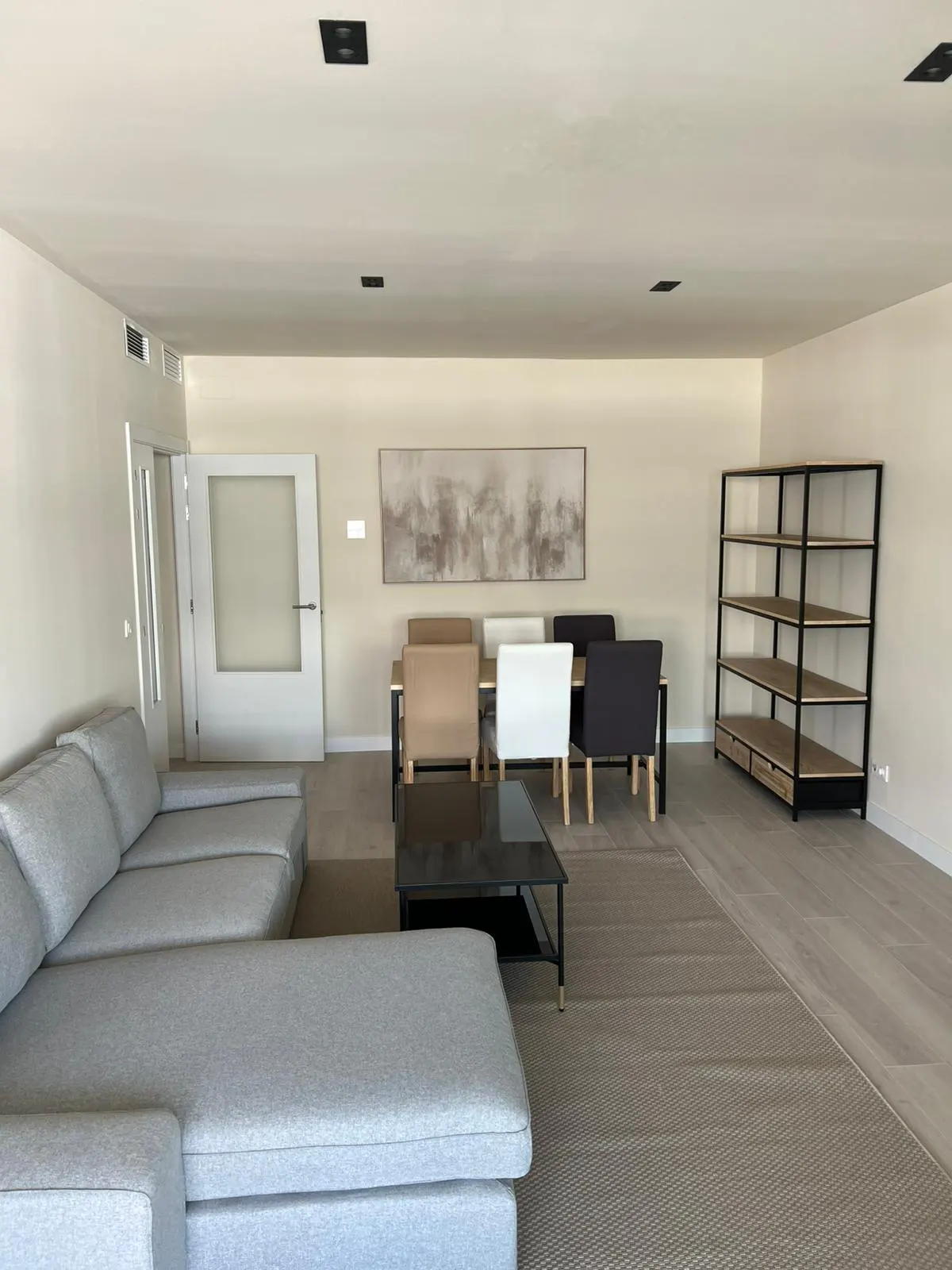 A bright, open-plan living and dining area with a gray sectional sofa, dining table with chairs, and a black metal shelving unit.