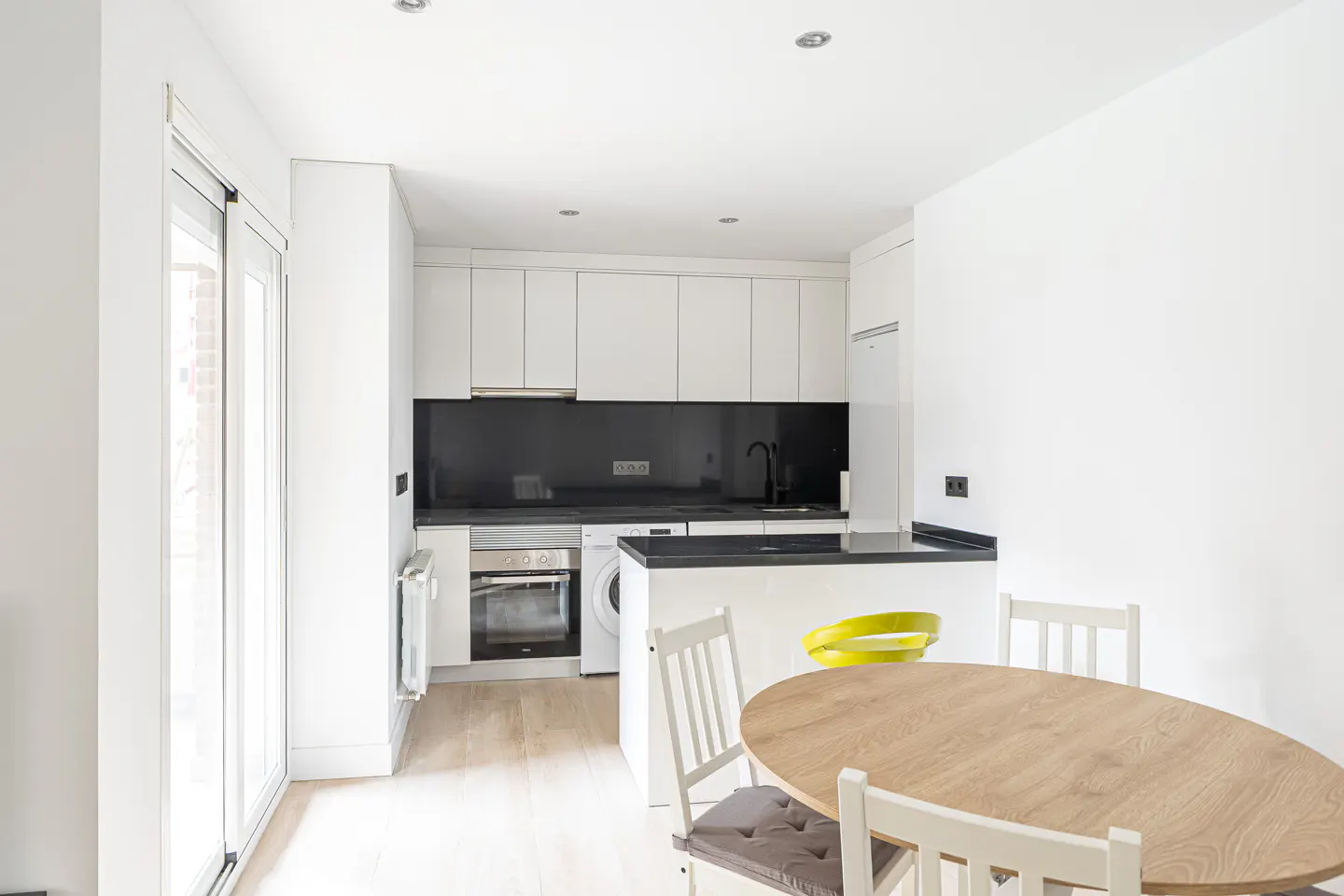 Bright, modern kitchen with white cabinets, black backsplash, and light wood floors. A round table with chairs sits in the foreground.