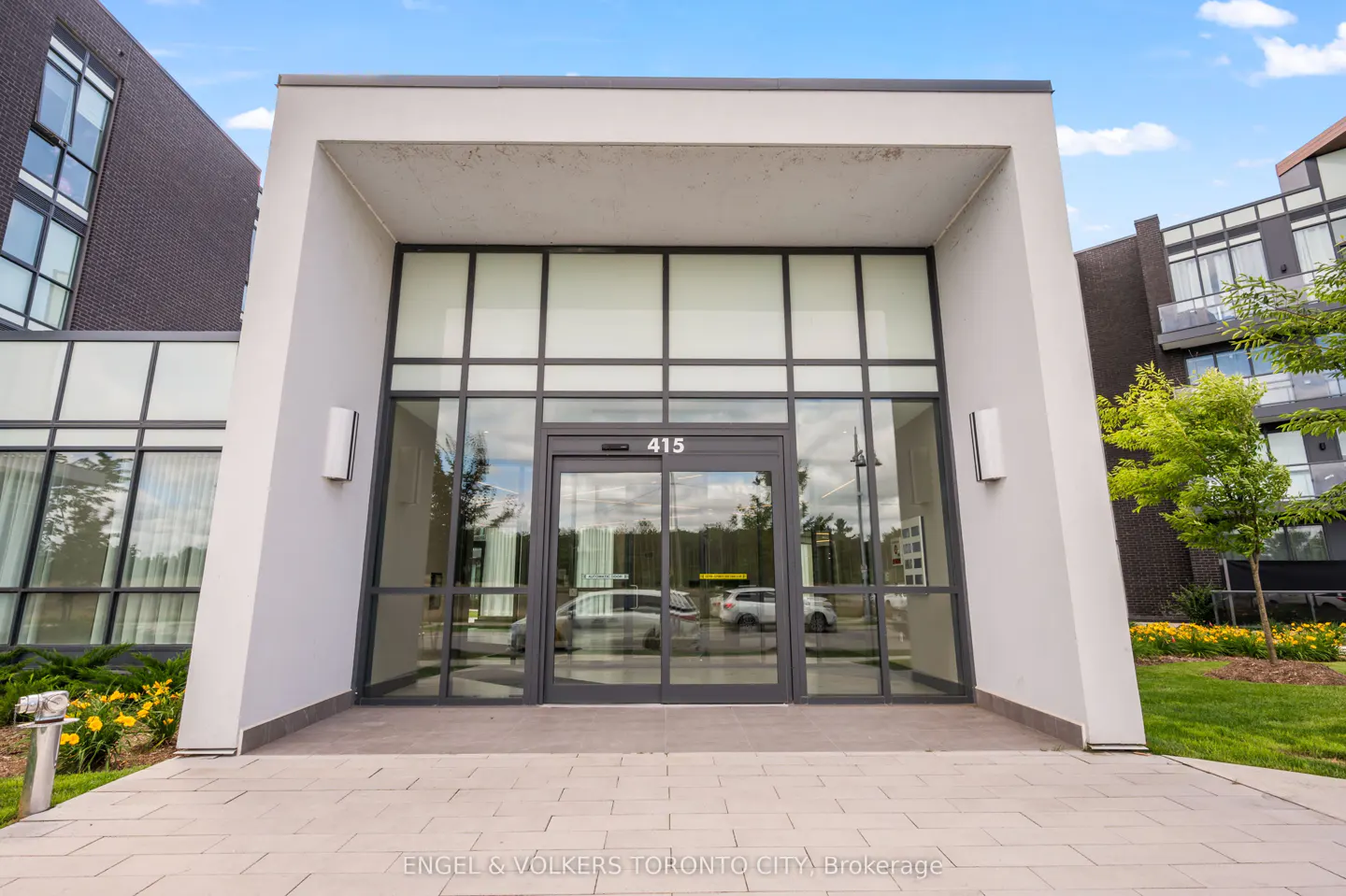 Modern building entrance with a white frame, glass doors labeled 415, and a paved walkway under a bright blue sky.