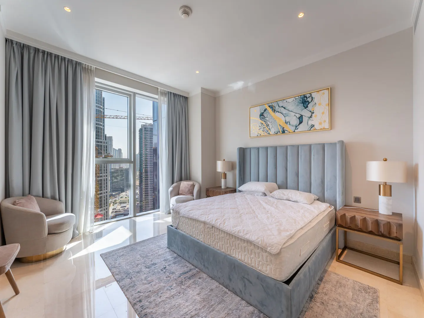 Bright bedroom with a blue bed, two chairs, and a city view from the window.