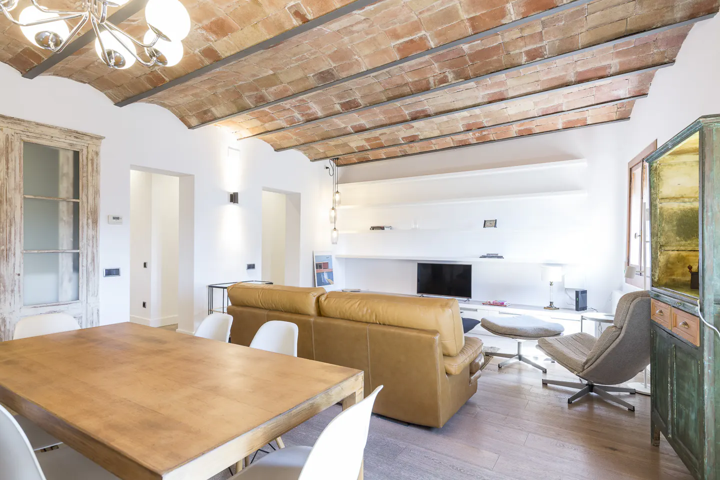 Bright living room with brick ceiling, wood floors, and white walls. A tan leather sofa faces a TV. A wooden table and chairs are in the foreground.