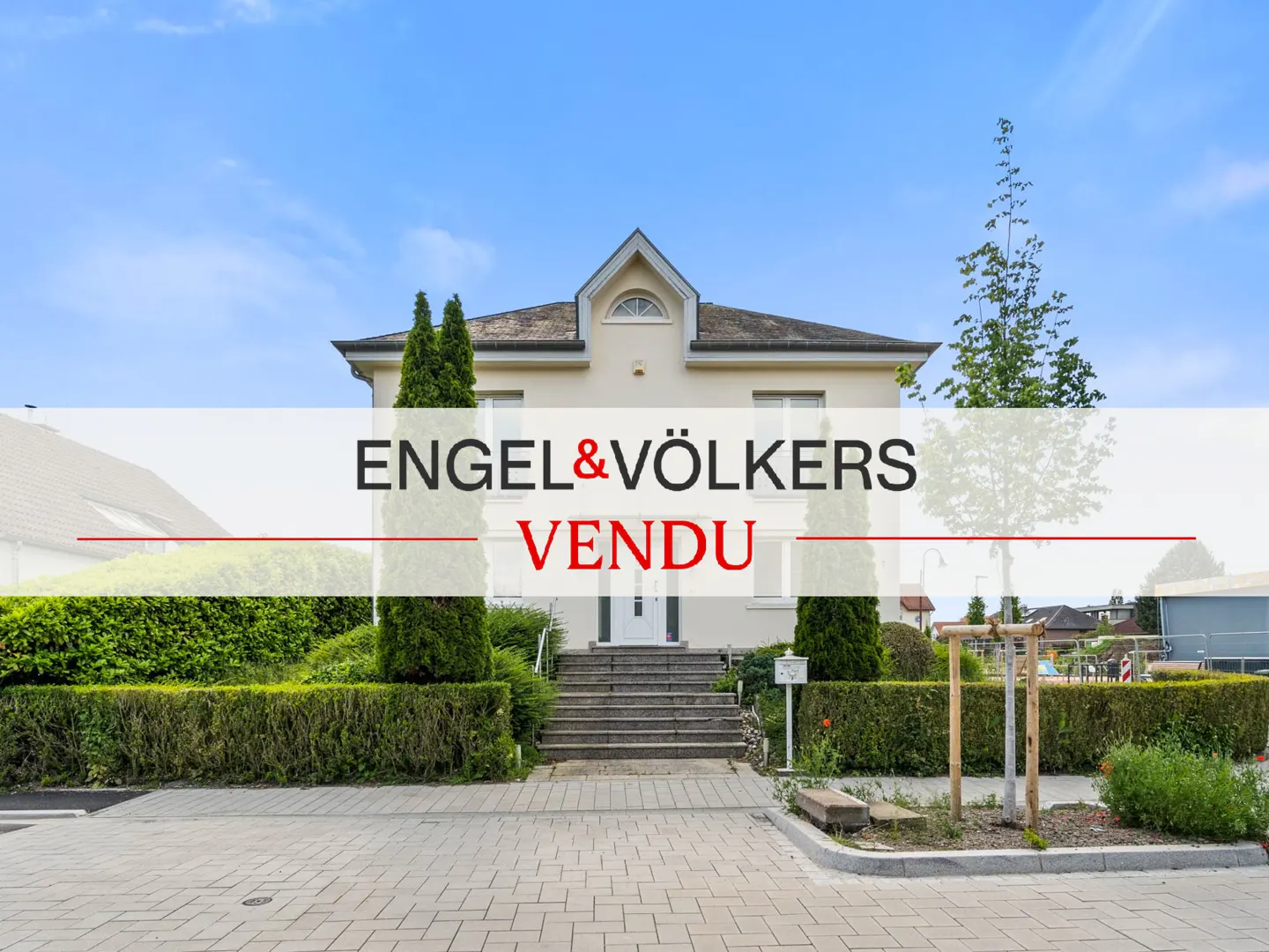 Sold! Beige two-story house with gray roof, green hedges, and stone steps leading to the front door under a blue sky. "Engel & Völkers Vendu" overlay.