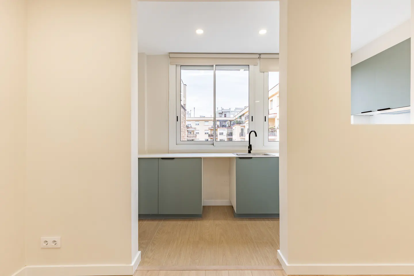 A view into a small kitchen area with light green cabinets, a black faucet, and a window overlooking city buildings. The walls are a light beige color.