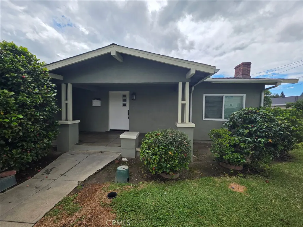 A single-story, gray house with a white front door and a brick chimney is surrounded by green bushes and grass.