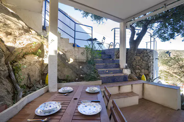 Outdoor dining area with a wooden table set for four, stone walls, and blue-tiled stairs leading to a terrace.
