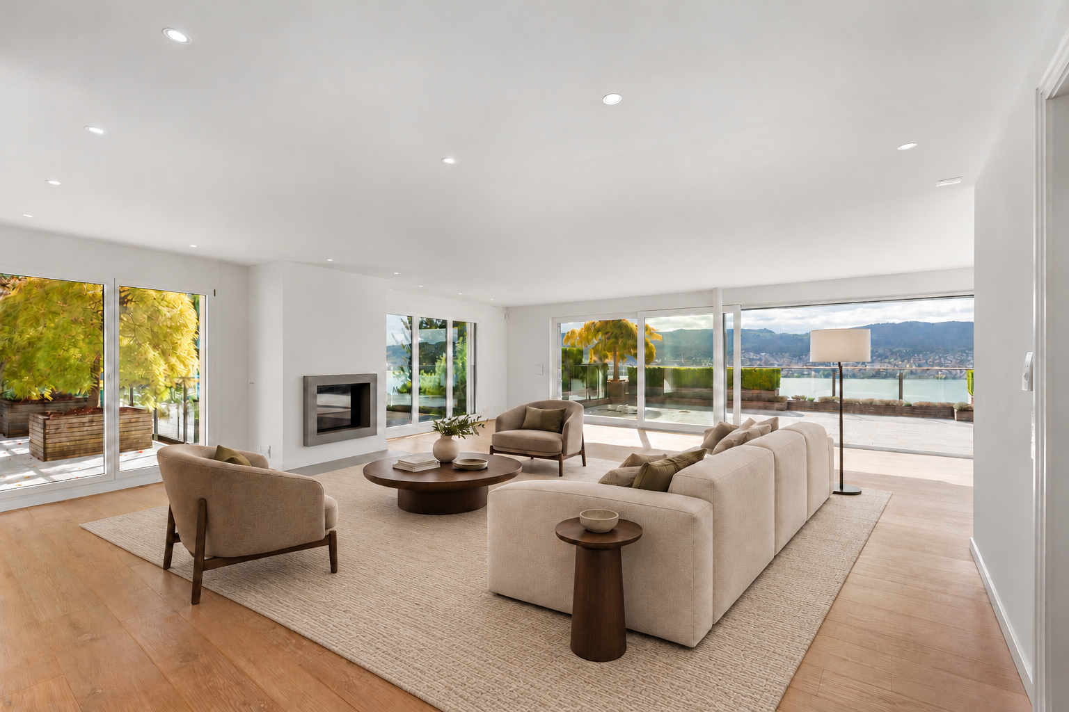 Modern living room with beige sofa, armchairs, round coffee table, fireplace, and large windows overlooking a scenic lake. [AI-generated text]