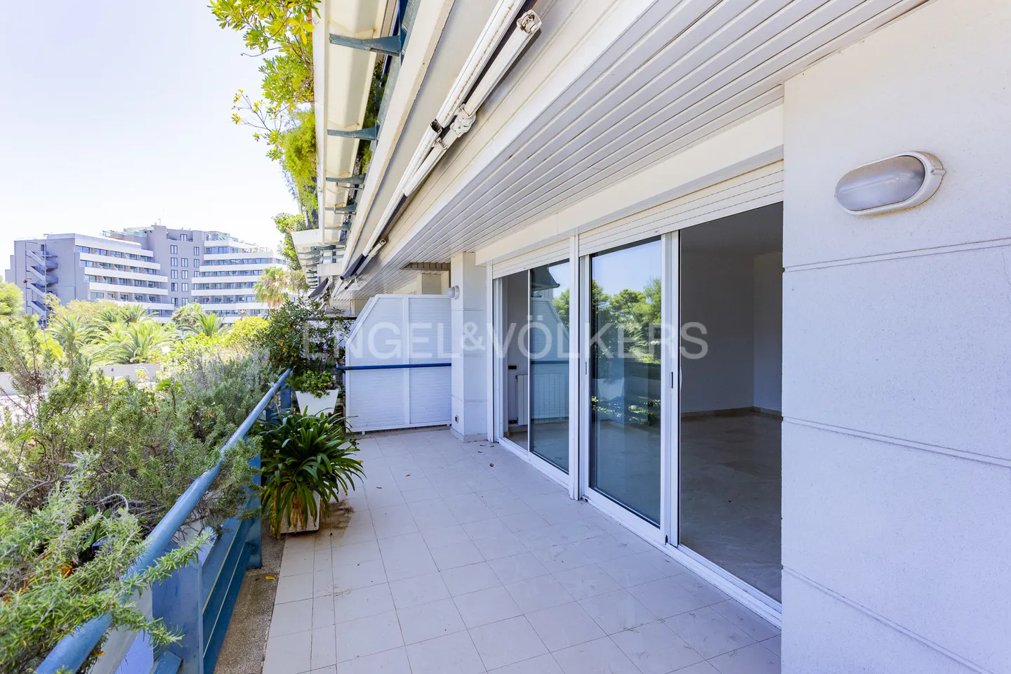 Wide balcony with tile floor, blue railing, and sliding glass doors. Plants line the railing, and a building is visible in the background.