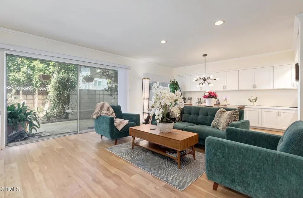 Living room with wood floors, green sofa and chairs, and a wood coffee table with white flowers. Sliding glass doors lead to a patio.