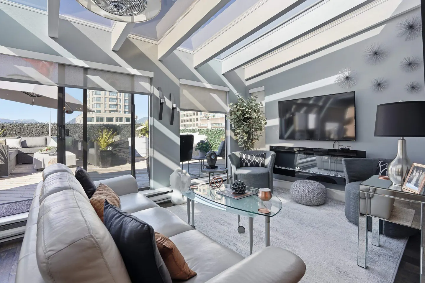 Bright living room with a white leather couch, glass coffee table, and a large TV. A patio is visible through sliding glass doors.