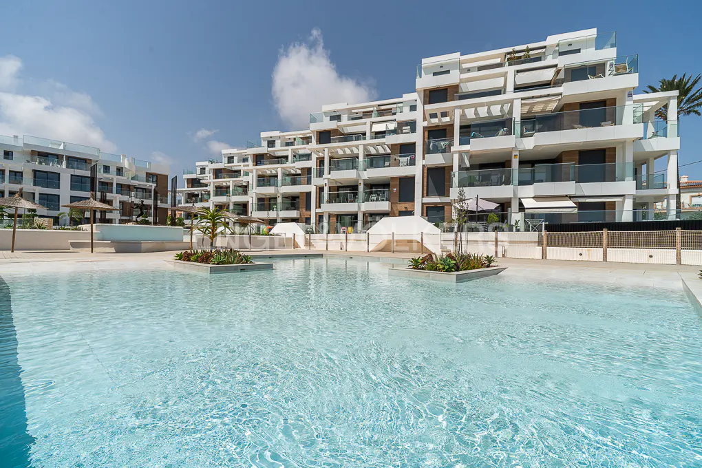 Exterior view of a modern, multi-story white apartment complex with a large, light blue swimming pool in the foreground.