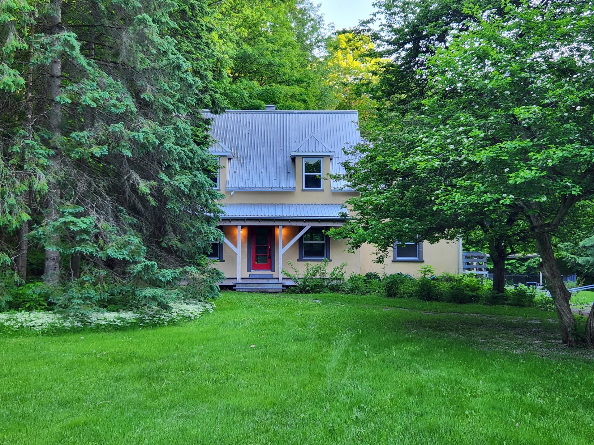 A two-story beige house with a metal roof and bright red front door, nestled in a lush green yard surrounded by trees.