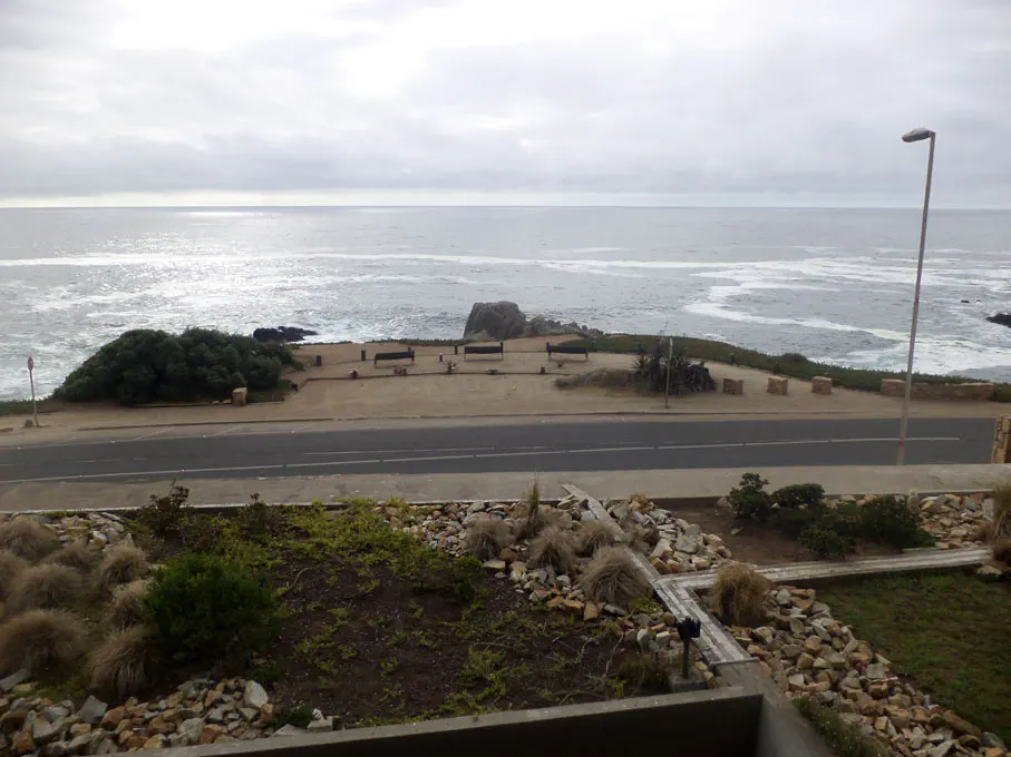 Ocean view from a balcony. A road separates the balcony from the ocean. Benches line the ocean view. Overcast sky.